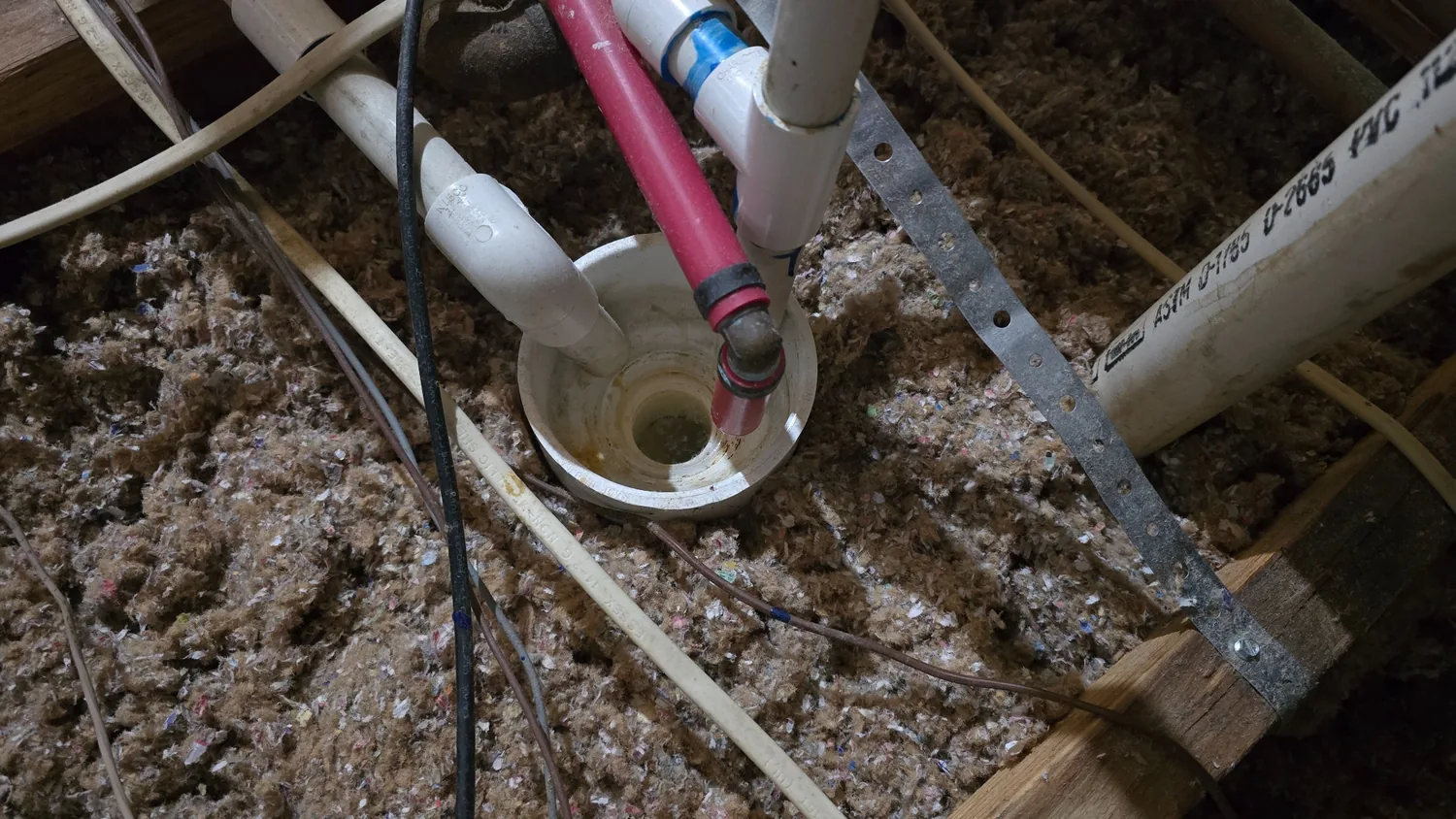 Actual open PVC condensate bowl receiving white and red inlet tubes over attic insulation, with a visible opening at its base